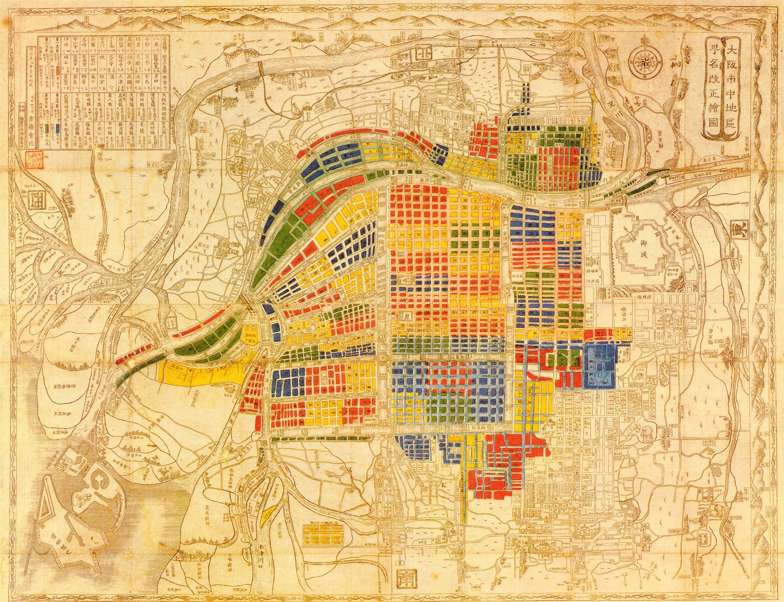 The blocks are washed flat red, blue, yellow and green. Everything else — the castle, the outlying villages, the paddy, the hills ruled along the top edge — stays in brown line, so the colour stops exactly where the new administrative districts stop. Under the wash is the Edo street plan, showing through in that same brown: same blocks, same canals, largely the same names.<br><br>The margins are modern. A compass rose beside the title at the top right, a table of distances at the top left, an inset of the river mouth at the bottom. Nothing here was surveyed for this sheet. It is an existing map of the city, coloured in to record a renaming.